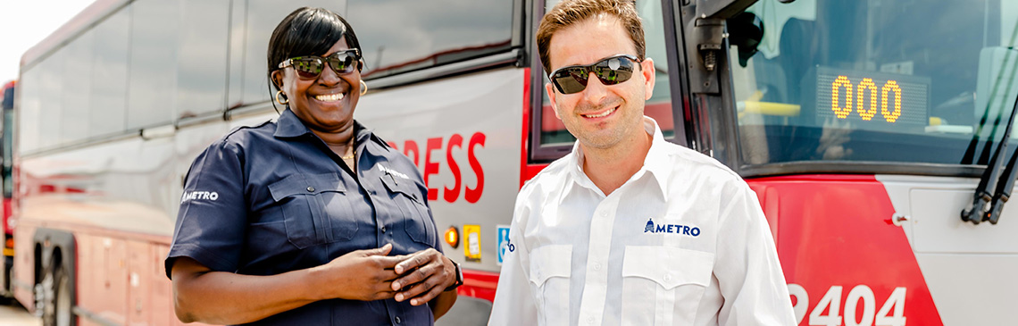 A female and a male MetroBus operator smiling while standing near a MetroExpess Bus