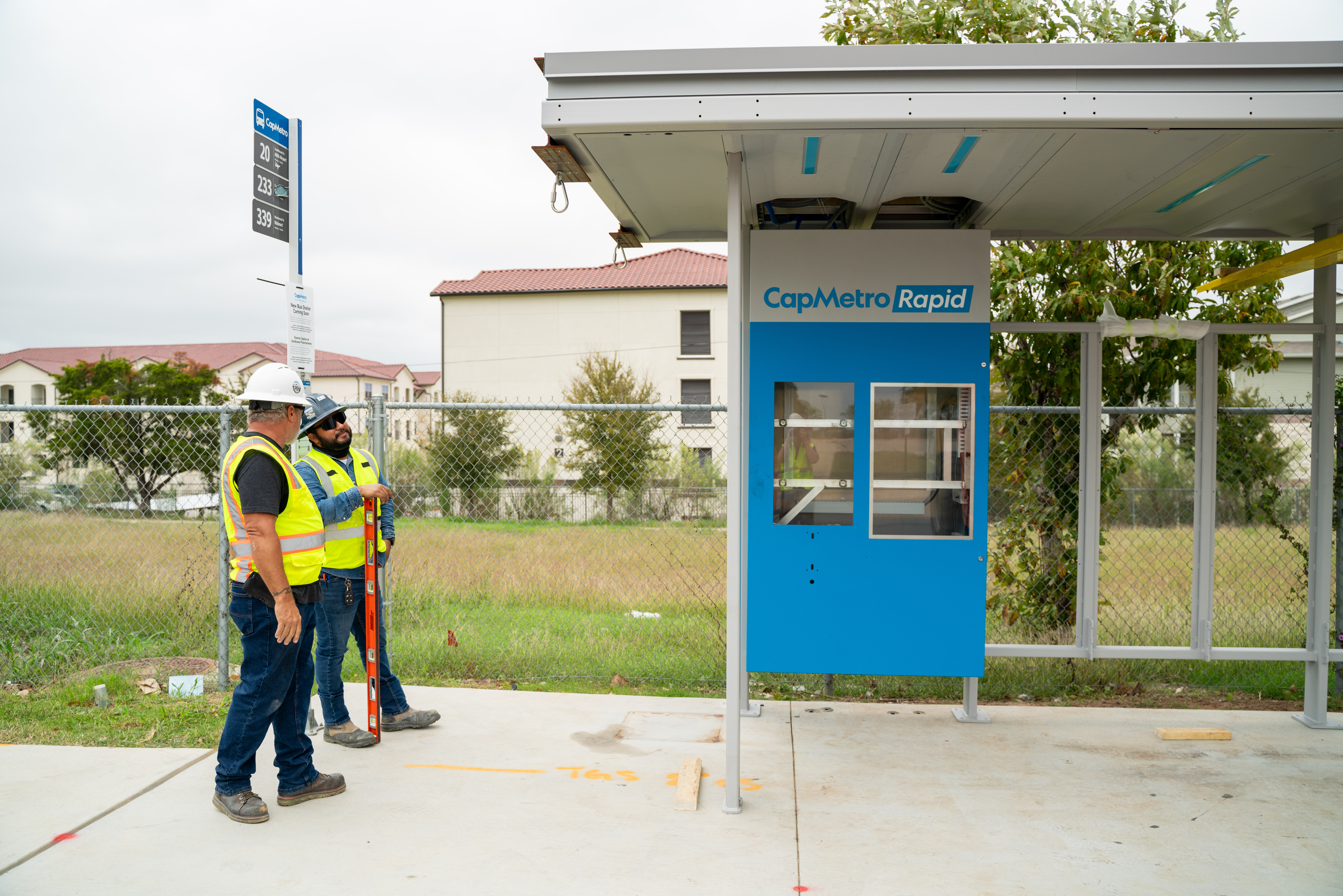 two construction workers checking their progress in front of a new, gray and blue CapMetro Rapid bus shelter