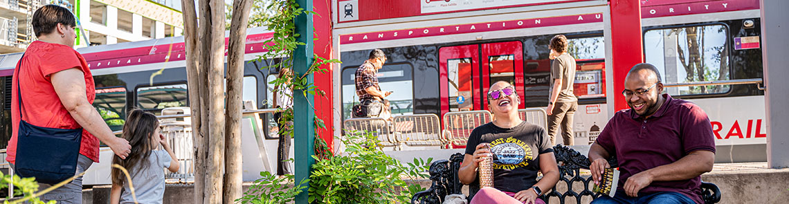 tod-hero-banner A diverse group of people – some walking, mingling, and sitting around a MetroRail station at Plaze Saltillo