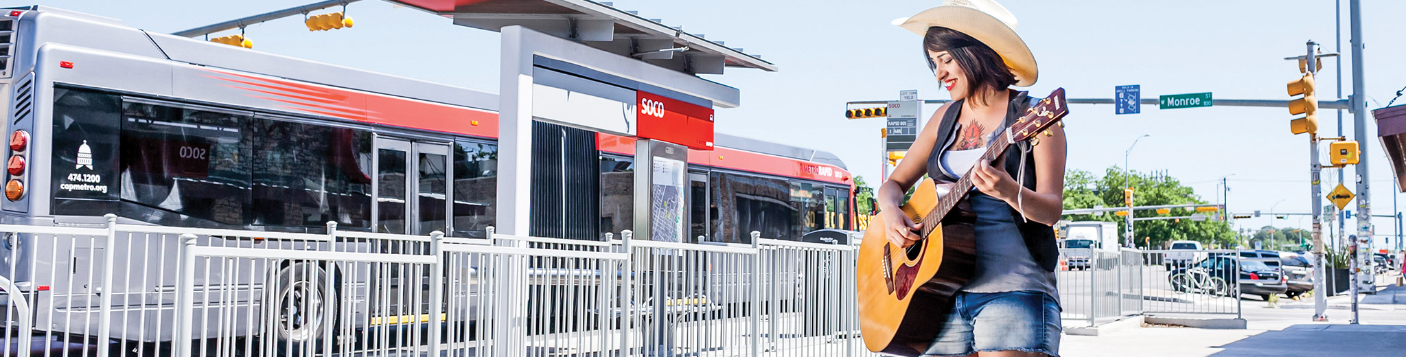 A large image of a young woman with jean shorts and a cowboy hat – playing a guitar at a MetroRapid bus station on a beautiful sunny day.